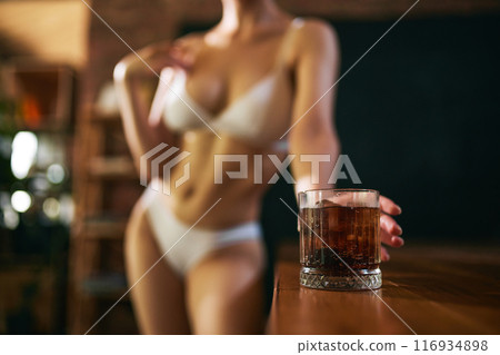 Woman in white lingerie stands near wooden bar, with glass of dark beverage in focus against blurred background. 116934898
