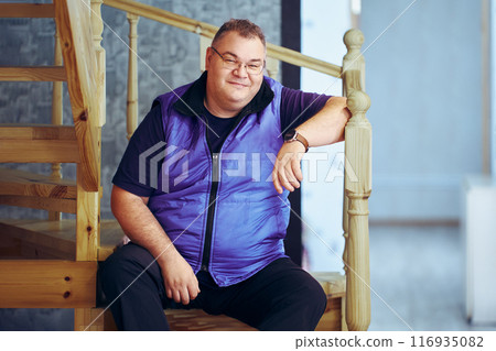Smug overweight white man in his 40s sits on step of wooden staircase. 116935082