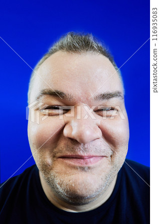 Malicious smile of 47 year old white man with an excess weight problem, his face distorted by wide-angle lens on blue background, closeup. Malicious smile of 47 year old white man with an excess weight problem, his face distorted by wide-angle lens on blue background, closeup. 116935083