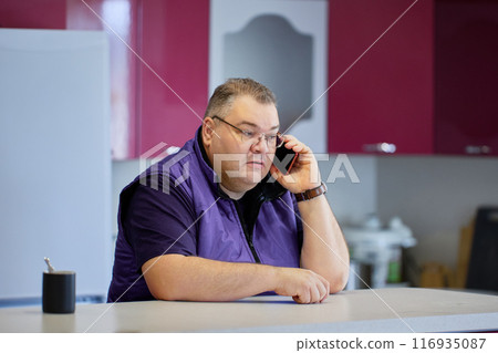 Large man listens attentively to his interlocutor on phone while in kitchen. 116935087