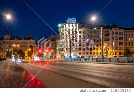 The Dancing House in Prague is illuminated by the city lights at dusk. The unique deconstructivist architecture stands out against the backdrop of traditional buildings. Light trails from passing cars 116935173