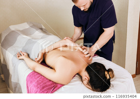Masseur massages woman shoulder area while she lies face down on massage table. Masseur massages woman shoulder area while she lies face down on massage table. 116935404