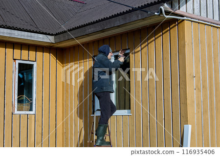 Exterior work on window openings, worker fixes metal window jambs on facade of boarded-up country house. Exterior work on window openings, worker fixes metal window jambs on facade of boarded-up country house. 116935406