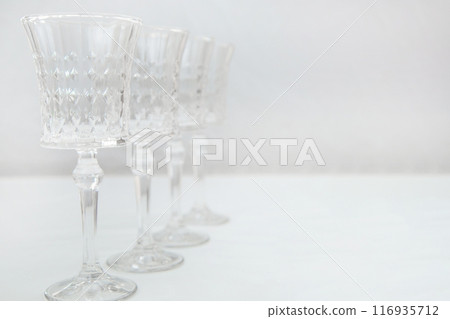 Crystal wine glasses on a long stem, with expanded edges and a faceted pattern stand in a row on a white background, the first one is in focus on the sides, the second ones stand behind it in focus 116935712