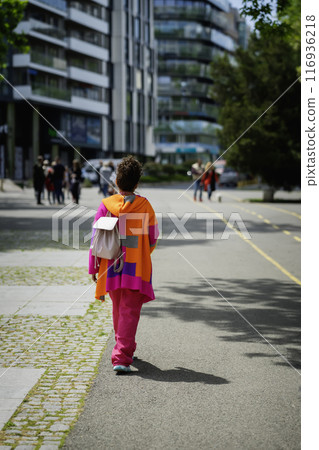 Girl in bright orange clothes and with a backpack walks along city street Girl in bright orange clothes and with a backpack walks along city street 116936218