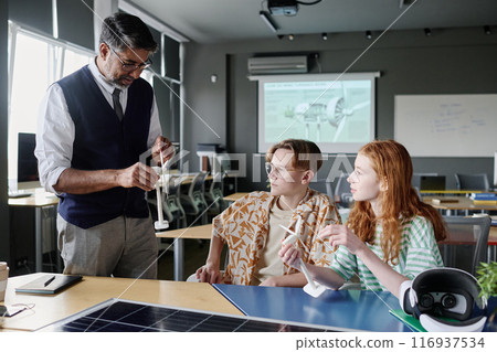 Medium long shot of mature teacher of Environmental Education showing teen students modern wind turbine model during lesson 116937534