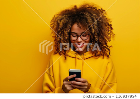 A modern woman plays a smartphone against an isolated yellow background. The concept of contemporary connectivity and integral part of daily life in a vibrant and energetic context. Generative AI. 116937954
