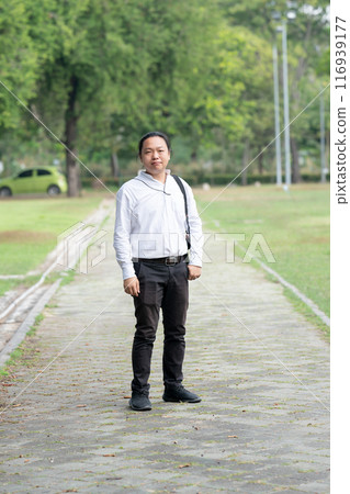 Asian freelance long hair man standing and posting on the walk way in the garden outdoor field with his camera beside him. Asian freelance long hair man standing and posting on the walk way in the garden outdoor field with his camera beside him. 116939177