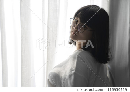 Asian Thai Chinese Woman with her black glasses stands in front of white curtain with the light from outside. It's a cute sexy shot image. 116939719