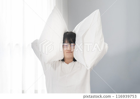 Asian woman raises pillow up to cover her ears and doesn't hear anything all around. 116939753