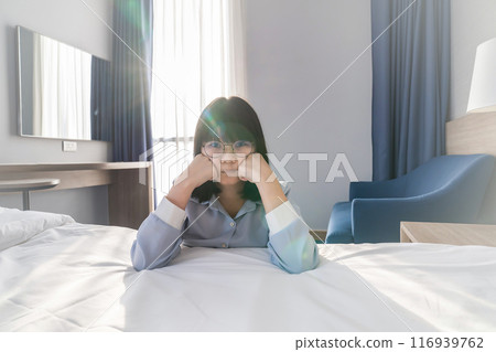 Pretty Asian Woman pucker up her lip in front of the bed in hotel bedroom, with the light from window curtain behind her. 116939762