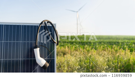 Close up of electric car charging plug with solar panel. Wind turbines in the background 116939859