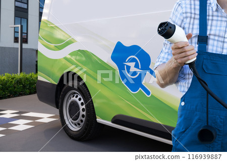 Man holds electric vehicle charging plug. Electric van in the background 116939887