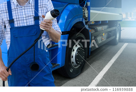Driver in uniform holds electric vehicle charging plug. Electric truck in the background 116939888