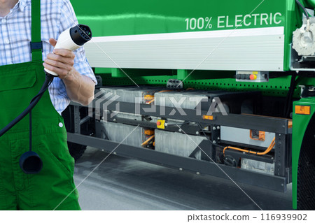 Driver in uniform holds electric vehicle charging plug. Electric truck in the background Driver in uniform holds electric vehicle charging plug. Electric truck in the background 116939902
