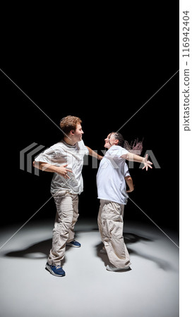 Stylish young man and woman in light matching outfits performing hip-hop, dancing against black studio background Stylish young man and woman in light matching outfits performing hip-hop, dancing against black studio background 116942404