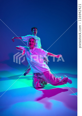 Talented young man and woman in casual sports clothes dancing hip-hop against purple studio background in neon light Talented young man and woman in casual sports clothes dancing hip-hop against purple studio background in neon light 116942421