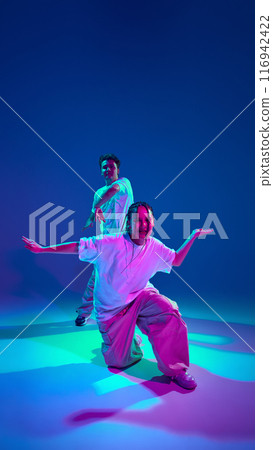 Artistic youth. Dynamic image of talented young girl and guy, hip-hop dancers in motion, performing against purple studio background in neon light Artistic youth. Dynamic image of talented young girl and guy, hip-hop dancers in motion, performing against purple studio background in neon light 116942422