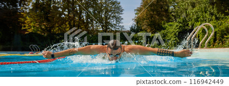 Swimmer, donning goggles, moves through pool with powerful butterfly stroke, surrounded by splashes and lush greenery in background. Swimmer, donning goggles, moves through pool with powerful butterfly stroke, surrounded by splashes and lush greenery in background. 116942449