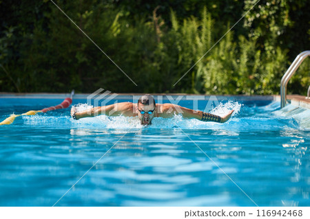 Focused and strong, swimmer with goggles executes butterfly stroke, propelling himself through water with precision. Pool outdoor. Focused and strong, swimmer with goggles executes butterfly stroke, propelling himself through water with precision. Pool outdoor. 116942468