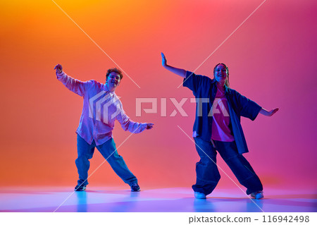 Dynamic image of young man and woman in casual oversized clothes dancing hip-hop against gradient studio background in neon light Dynamic image of young man and woman in casual oversized clothes dancing hip-hop against gradient studio background in neon light 116942498