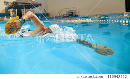 Swimmer wearing white cap, and yellow paddles trains in clear blue pool outside, perfecting his freestyle stroke on bright sunny day. 116942512
