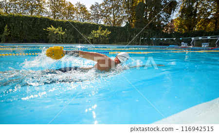 Athletic man wearing white swim cap, and goggles swims freestyle, utilizing yellow paddle to enhance his performance in sunny pool. 116942515