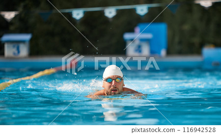 Determined swimmer in white cap and goggles glides through pool, focused on his training against blurred sunny outdoor background. 116942528