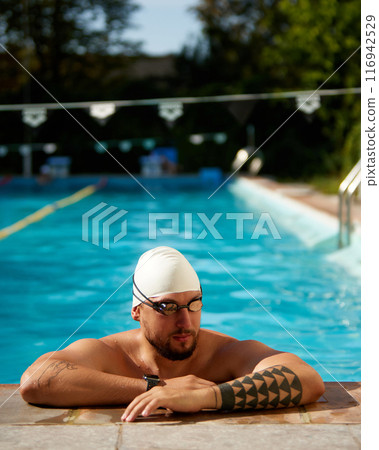 Swimmer in white swim cap rests at pool's edge, wearing goggles and looking thoughtful against sunny outdoor backdrop. 116942529