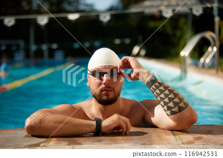 Young man, swimmer in swimming hat and goggles leaned on edge of pool and confidently looks at camera ready to swimming. 116942531