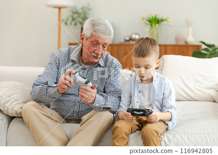 Happy family. Old senior man grandfather boy grandson playing video game with joysticks at home. Smiling grandparent and child using gamepads for video game. Older generation modern tech usage 116942805