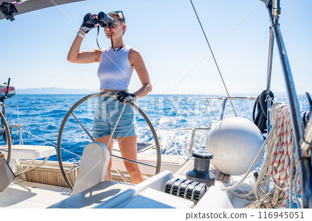 Young female captain on the yacht looking through binoculars during sailing boat control. Travel and active life. Yachting Young female captain on the yacht looking through binoculars during sailing boat control. Travel and active life. Yachting 116945051