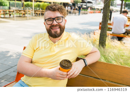 Millennial smiling bearded man in yellow polo t-shirt eyeglasses sitting on bench drink coffee have breakfast rest relax in sunshine spring green city park outdoors on nature. Urban leisure concept Millennial smiling bearded man in yellow polo t-shirt eyeglasses sitting on bench drink coffee have breakfast rest relax in sunshine spring green city park outdoors on nature. Urban leisure concept 116946319