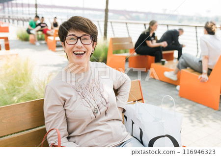 Outdoor portrait of beautiful and elegant laughing middle age 55 - 60 year old woman wearing long sleeve top sitting on bench on the street . Copy space and empty place for text advertising Outdoor portrait of beautiful and elegant laughing middle age 55 - 60 year old woman wearing long sleeve top sitting on bench on the street . Copy space and empty place for text advertising 116946323