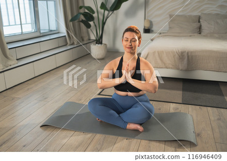 Yoga mindfulness meditation. Woman practicing yoga in living room at home. Plus size female sitting in lotus pose meditating smiling relaxing indoor. Girl doing breathing practice. Yoga at home Yoga mindfulness meditation. Woman practicing yoga in living room at home. Plus size female sitting in lotus pose meditating smiling relaxing indoor. Girl doing breathing practice. Yoga at home 116946409