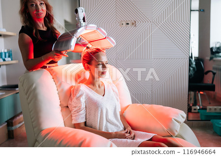 Side view of pretty blonde female having LED light hair photodynamic therapy treatment at modern medical clinic. Cosmetician specialist adjusting innovation device for PDT therapy procedure Side view of pretty blonde female having LED light hair photodynamic therapy treatment at modern medical clinic. Cosmetician specialist adjusting innovation device for PDT therapy procedure 116946664