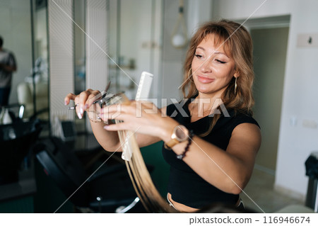 Closeup portrait of female hairdresser making stylish haircut with professional scissors to young woman client with long straight blonde hair. Hairstylist cutting lady hair with barber scissors. 116946674