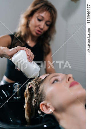 Vertical shot of professional hairstylist foaming shampoo on long blonde hair of beautiful female client. Skilled hairdresser washing head of young woman in beauty hairdressing salon. 116946677