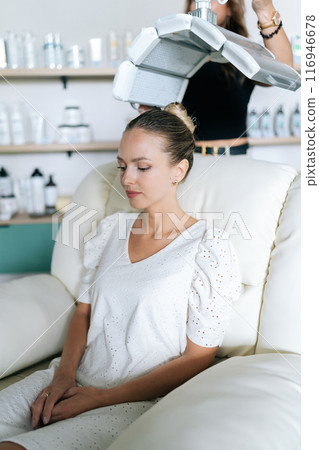 Vertical portrait of woman client with blonde hair having LED light hair PDT treatment at luxury aesthetic clinic. Female specialist adjusting innovation device for photodynamic therapy procedure 116946678