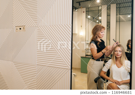 Professional woman hairstylist applying peeling purifying and soothing cosmetics product to scalp using cotton swabs. Reflection on mirror of blonde female having professional hair beauty treatment. Professional woman hairstylist applying peeling purifying and soothing cosmetics product to scalp using cotton swabs. Reflection on mirror of blonde female having professional hair beauty treatment. 116946681