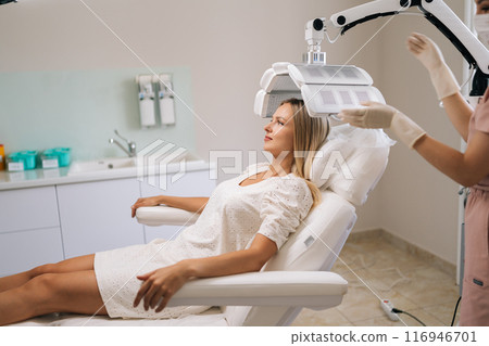 Side view of beautician specialist connecting and adjusting innovation device for photodynamic therapy procedure during hair treatment at modern medical clinic. Concept of health checkup, treatment. 116946701