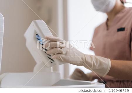 Close-up cropped shot of unrecognizable dermatologist in rubber gloves setting modern device in medical clinic. Closeup hands of female doctor adjusting apparatus, touching touch screen with finger. 116946739
