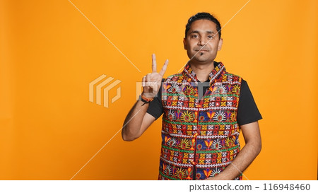 Portrait of upbeat man crossing fingers to make wish, hoping for good luck, isolated over studio background. Jolly indian person doing peace victory hand sign gesture, camera B 116948460