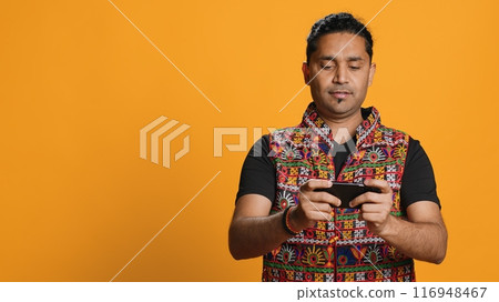 Indian man playing videogames on cellphone in landscape mode, enjoying leisure time. Gamer enjoying game on mobile phone, having fun, isolated over studio background, camera A 116948467