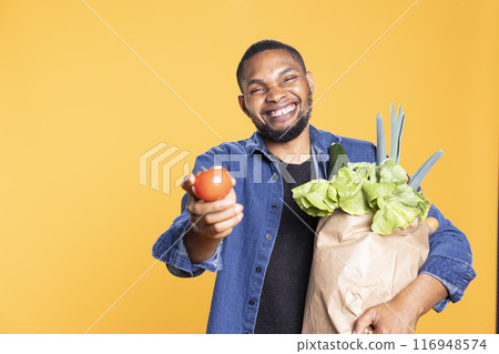 Portrait of young man presenting natural delicious tomato in studio, holding a paper bag full of natural eco friendly bio food. Happy cool guy looks at organic delicious veggies in studio. 116948574