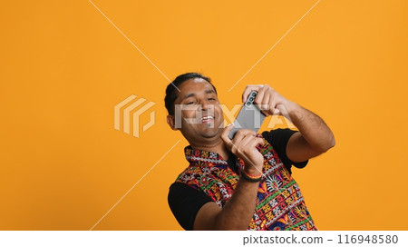 Cheerful Indian person in traditional clothing entertained by videogames on smartphone. Joyous player enjoying game on phone, having fun online, studio backdrop, camera A 116948580