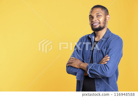 Portrait of confident african american person posing in studio, standing with arms crossed against yellow background. Enthusiastic relaxed guy smiling in front of the camera, masculine figure. 116948588