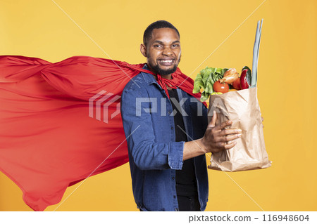 Superhero model with red cape posing with a bag of bio groceries, promoting sustainable lifestyle and locally grown veggies. Young person showcasing zero waste concept in studio. 116948604