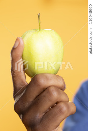 African american guy holding a green apple against yellow background, presenting local ethically sourced fresh produce in front of the camera. Zero waste enthusiast shows ripe fruits. Close up. 116948660