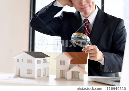 A worried man in a suit looking at a model house through a magnifying glass 116949810
